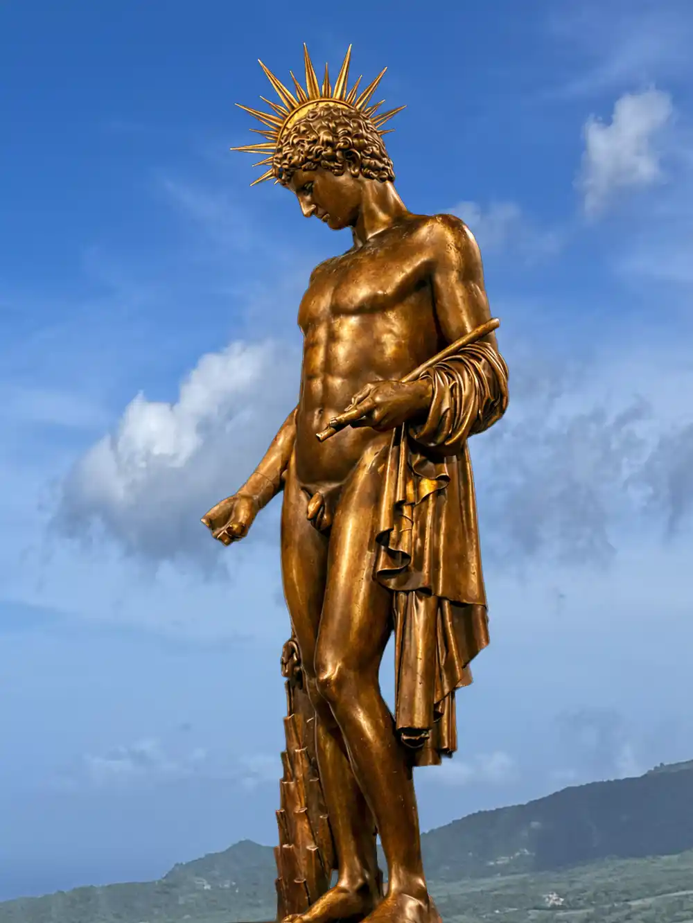Eine goldene Statue einer stehenden männlichen Figur mit Strahlenkranz vor einem blauen Himmel und Bergen.