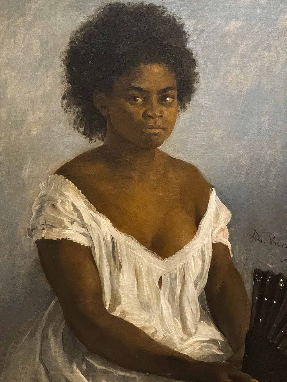 Eine Frau mit lockigem Haar in einem weißen Kleid sitzt vor einem schlichten Hintergrund und schaut direkt nach vorne.