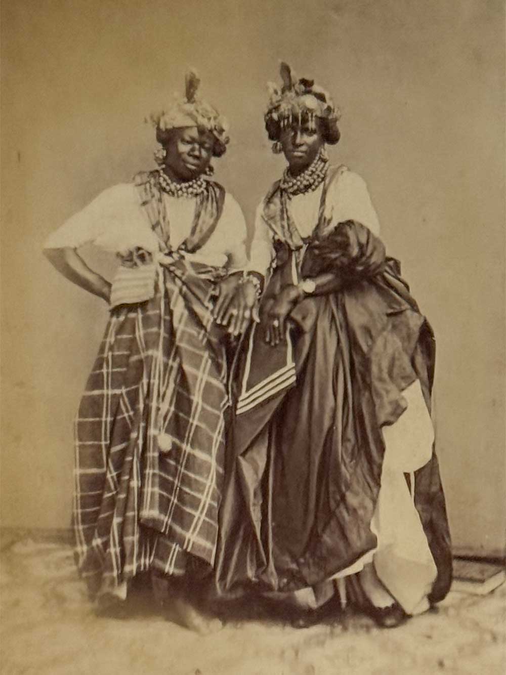 Auf einer alten Sepia-Fotografie stehen zwei Frauen in traditioneller Kleidung und mit Schmuck nebeneinander und schauen in die Kamera.