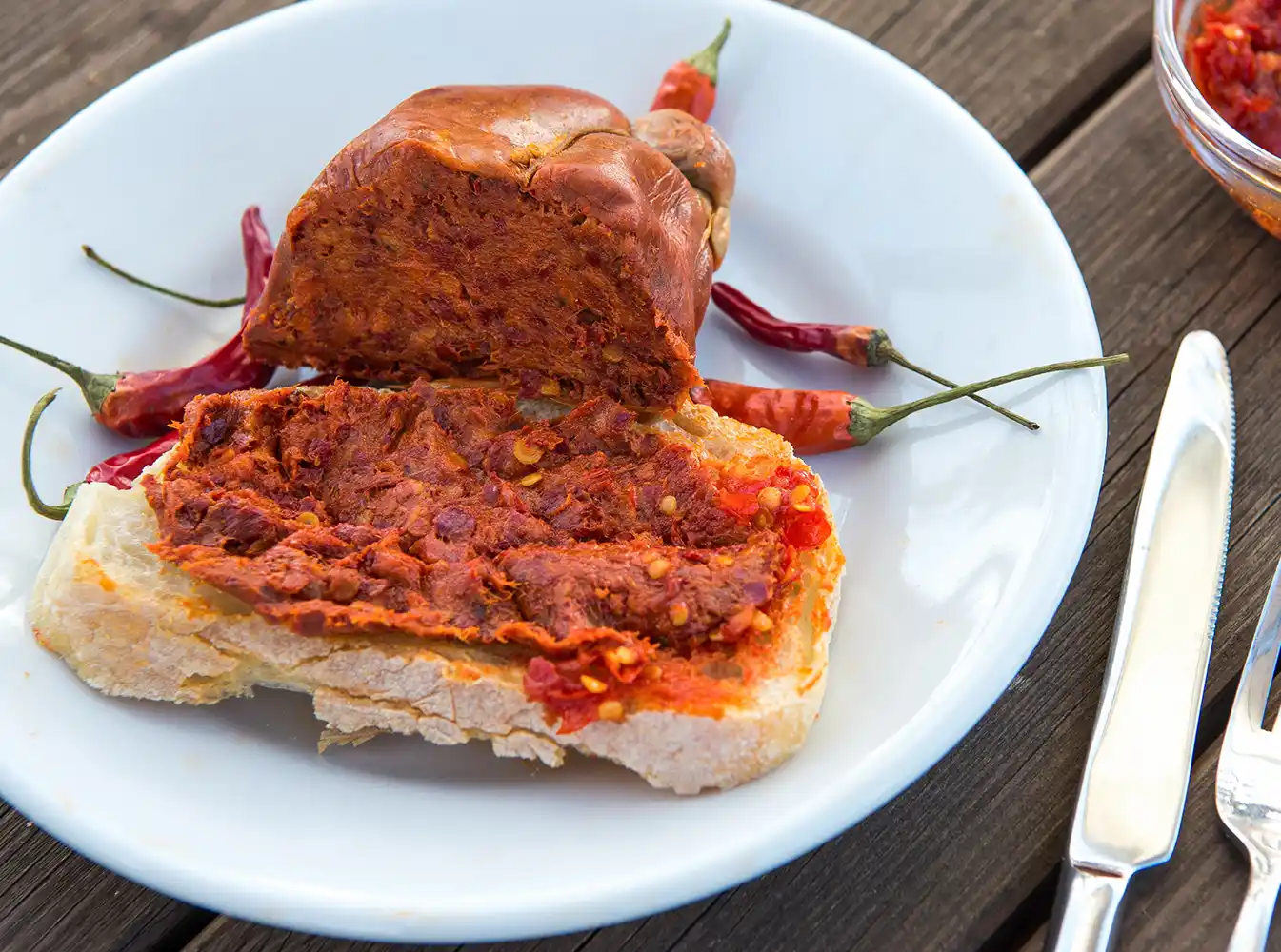 Brot mit würzigem Aufstrich, Wurstscheiben und roten Chilischoten auf einem weißen Teller mit Besteck.