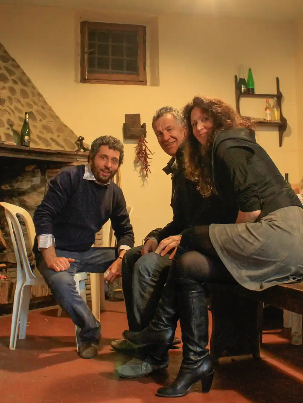 Drei Erwachsene posieren und lächeln in einem gemütlichen Raum neben einem Steinkamin und Holzregalen mit Flaschen.