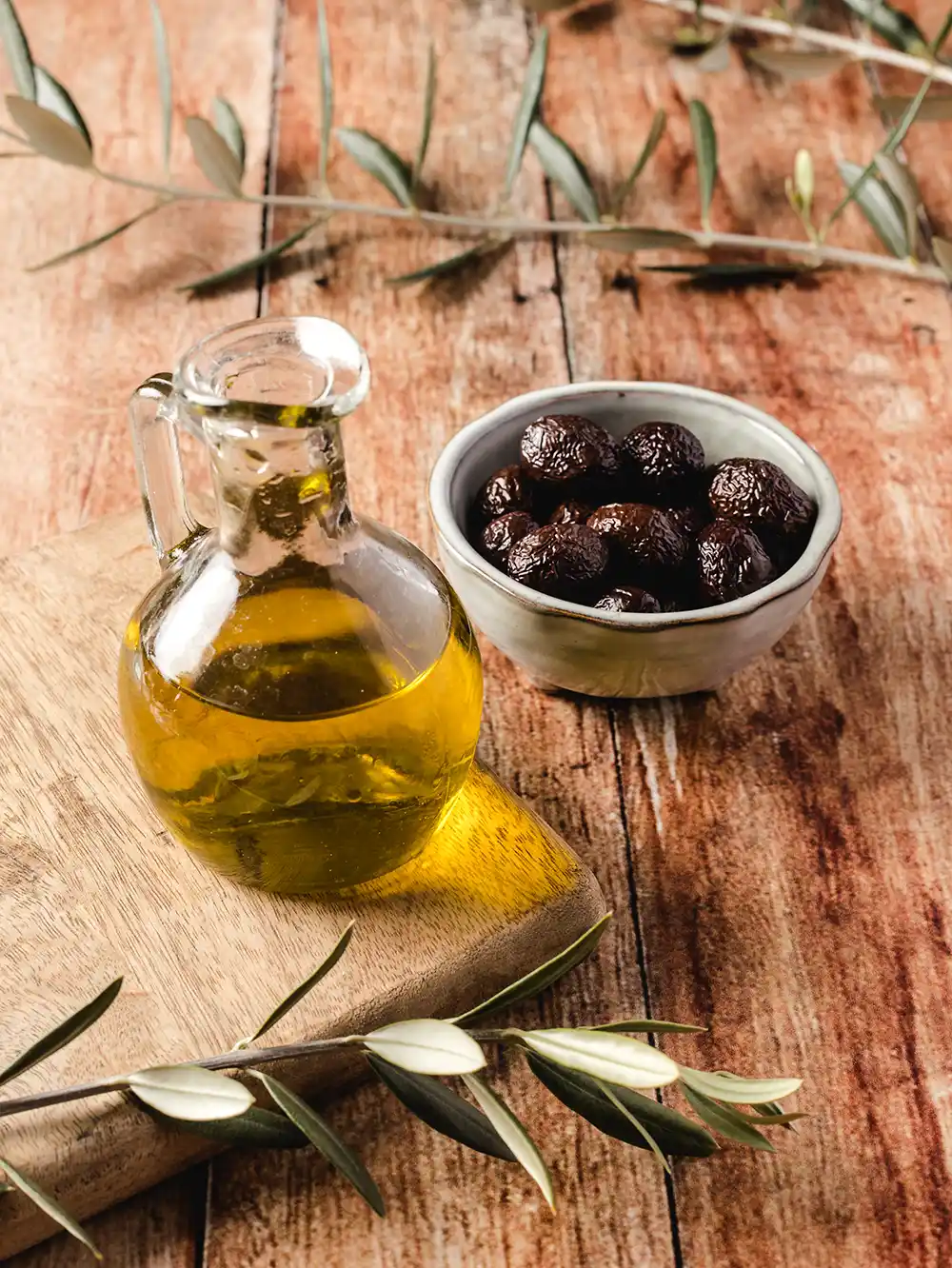 Ein Glaskrug mit Olivenöl und eine Schale mit schwarzen Oliven auf einem Holztisch mit Olivenzweigen.