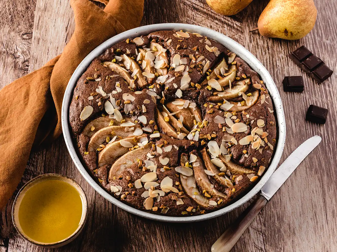 Schokoladenbirnenkuchen mit geschnittenen Birnen und Mandeln auf einem Holztisch, daneben ein Messer, Honig und Birnen.