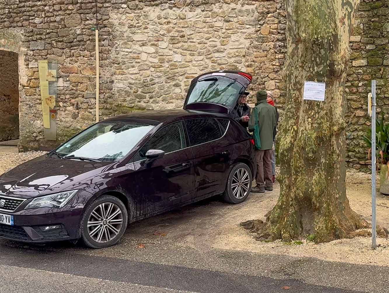 Zwei Personen stehen an einem bewölkten Tag an einem offenen Kofferraum neben einem Baum und einer Steinmauer.