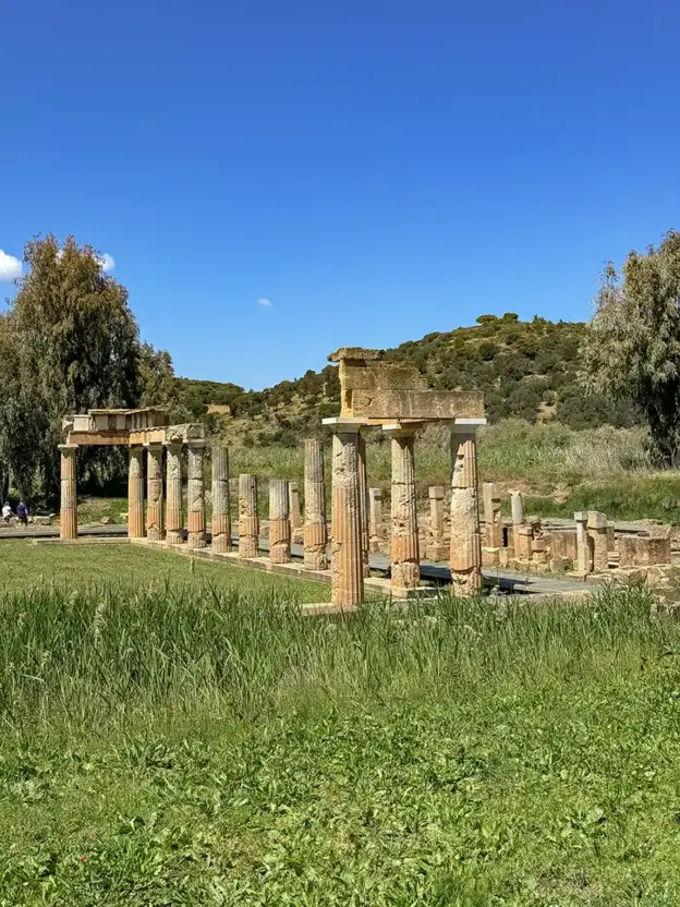 Alte Steinsäulen und Ruinen stehen in einem grasbewachsenen Feld mit Bäumen und Hügeln im Hintergrund unter einem blauen Himmel.