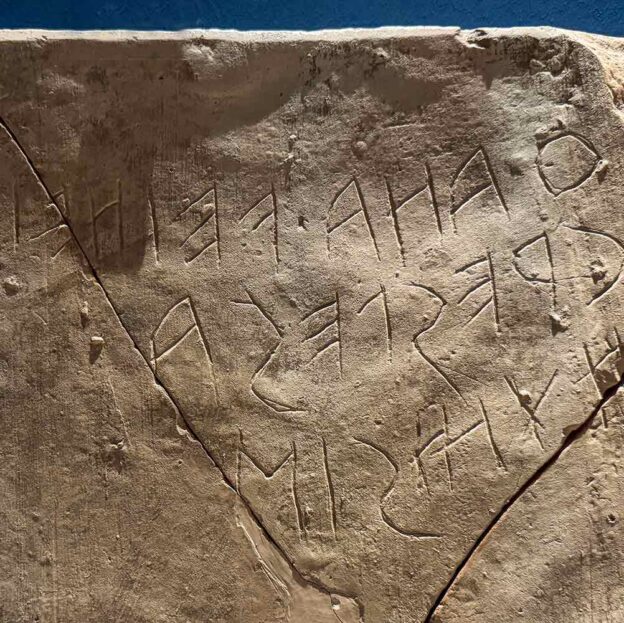 Alte Steintafel mit teilweise zerbrochener Inschrift in einem alten Alphabet vor blauem Hintergrund.