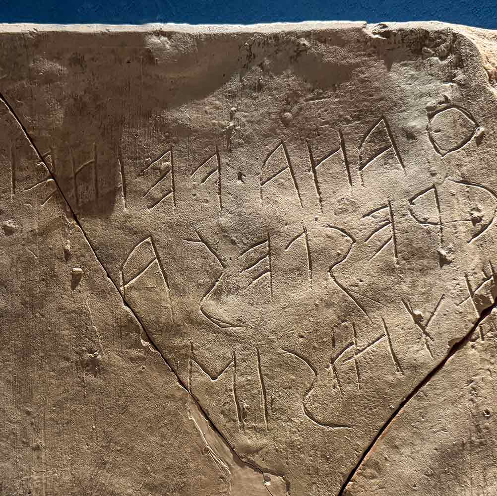 Alte Steintafel mit teilweise zerbrochener Inschrift in einem alten Alphabet vor blauem Hintergrund.