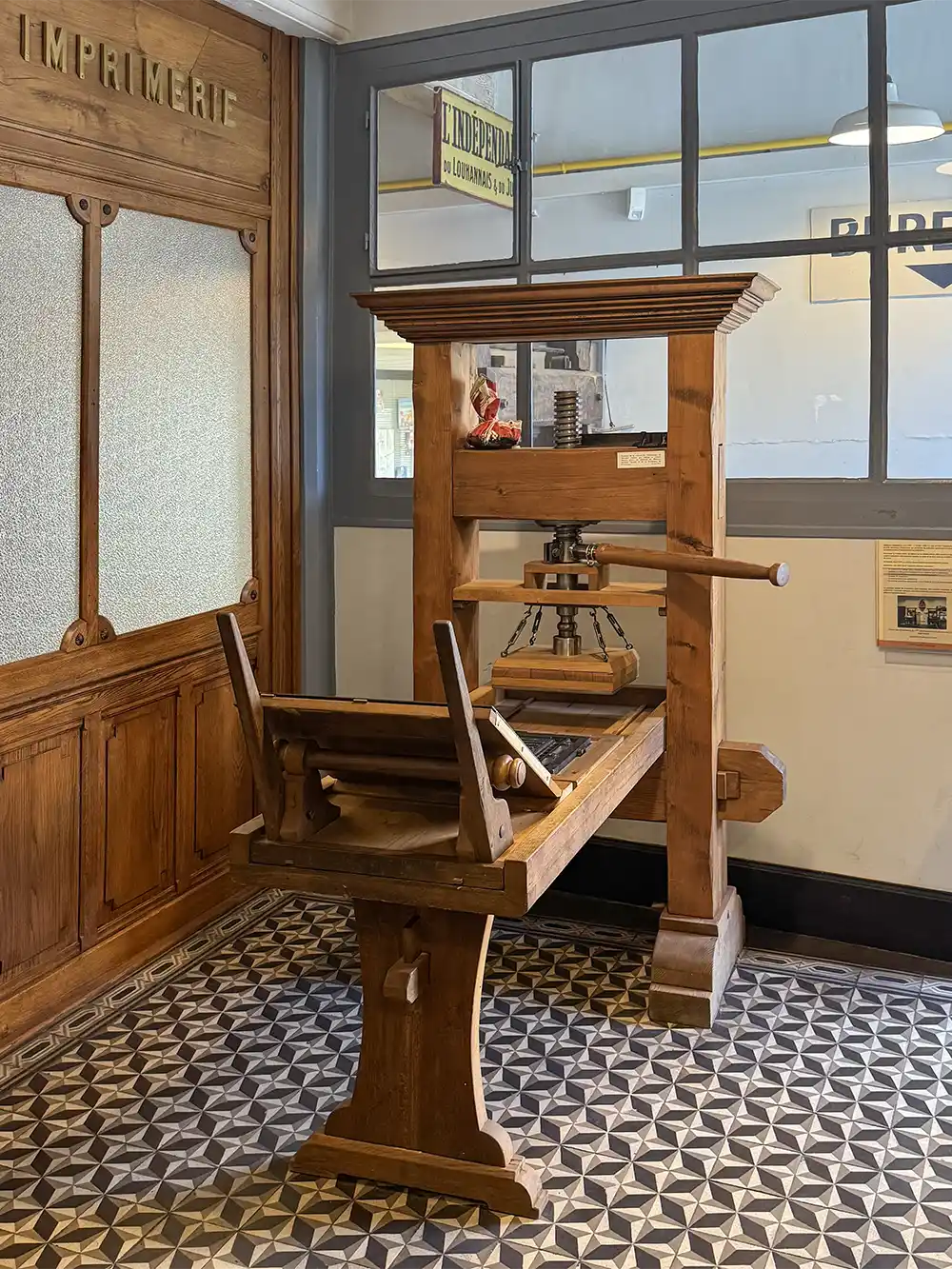 Eine alte Druckerpresse aus Holz in einem Raum mit gemustertem Fliesenboden und Glasfenstern.