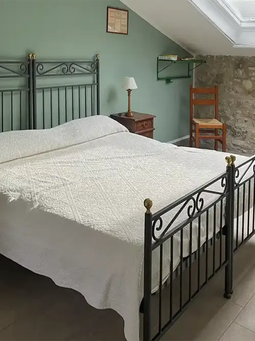 Gemütliches Schlafzimmer mit zwei Eisenbetten, weißem Bettzeug, Steinmauer, Holzmöbeln und einem Dachfenster.