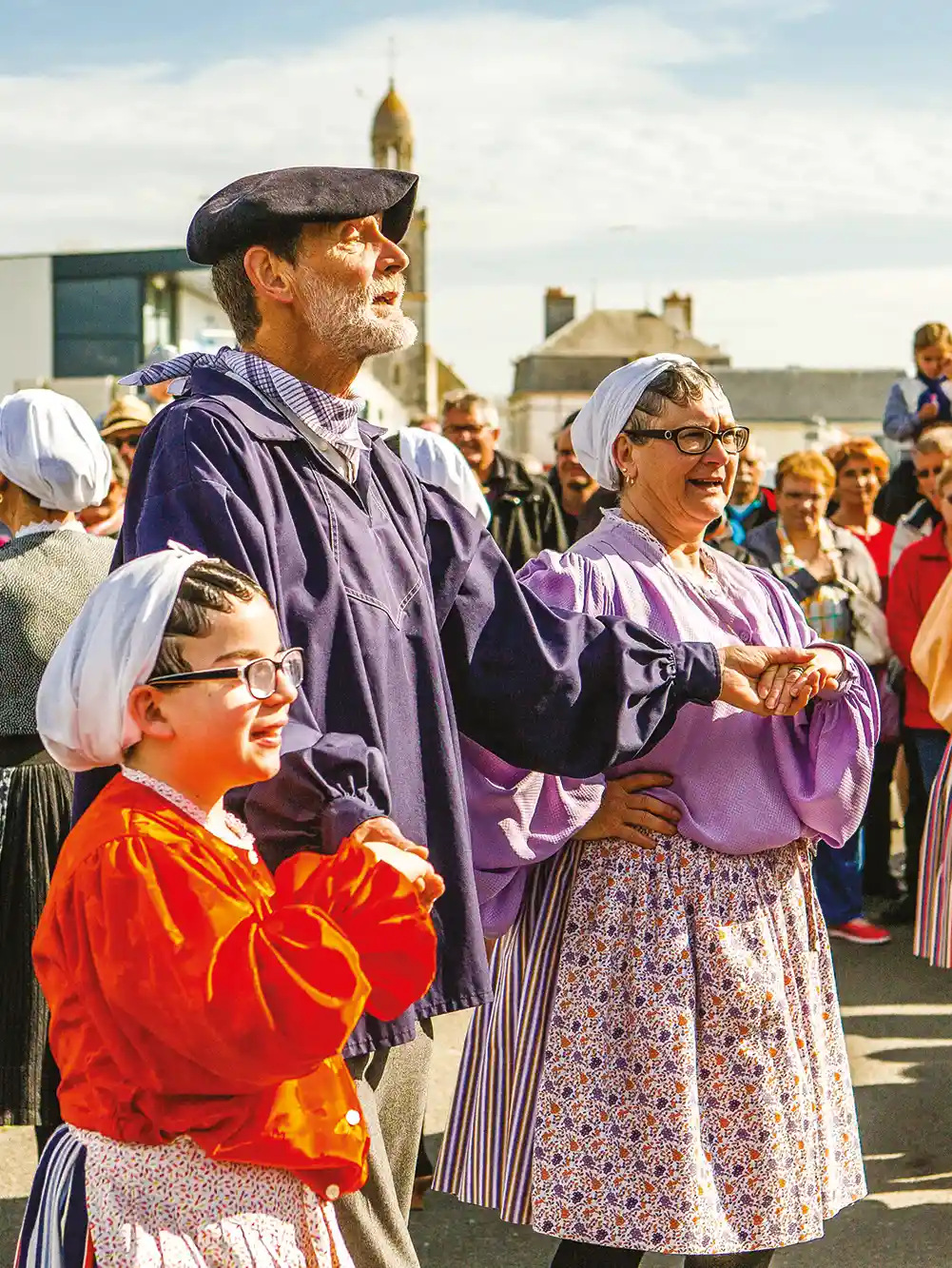Erwachsene und ein Kind in traditioneller Kleidung tanzen während eines Festes unter freiem Himmel, beobachtet von einer Menschenmenge.