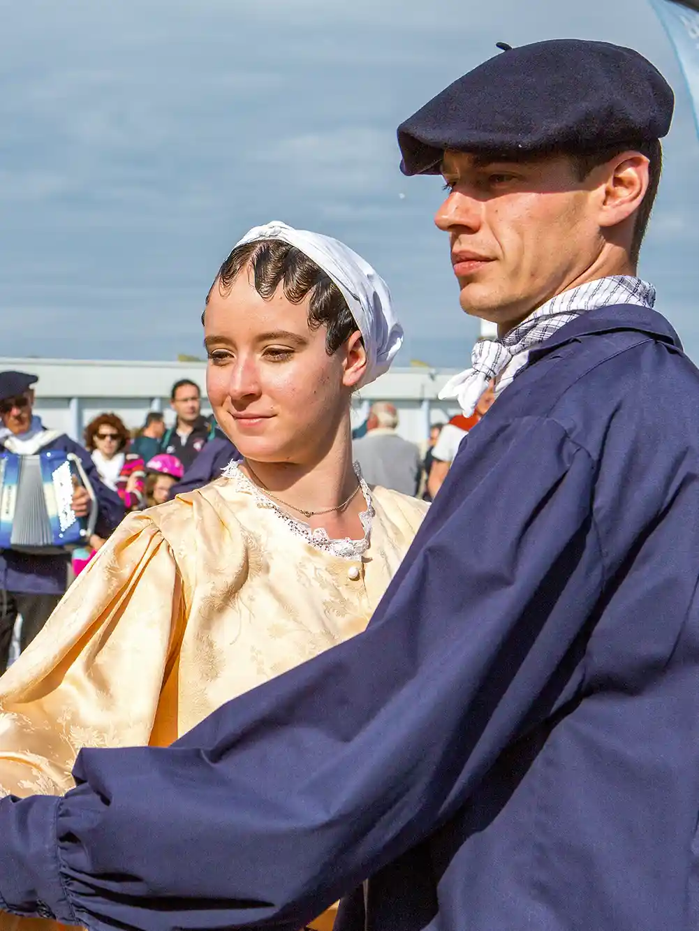 Ein Mann und eine Frau in traditioneller französischer Tracht tanzen während eines Festes im Freien, mit Menschen im Hintergrund.