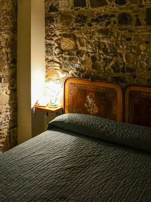 Ein Bett mit einer dunklen Tagesdecke neben einem hölzernen Kopfteil und einer brennenden Lampe auf einem kleinen Tisch vor einer Steinwand.