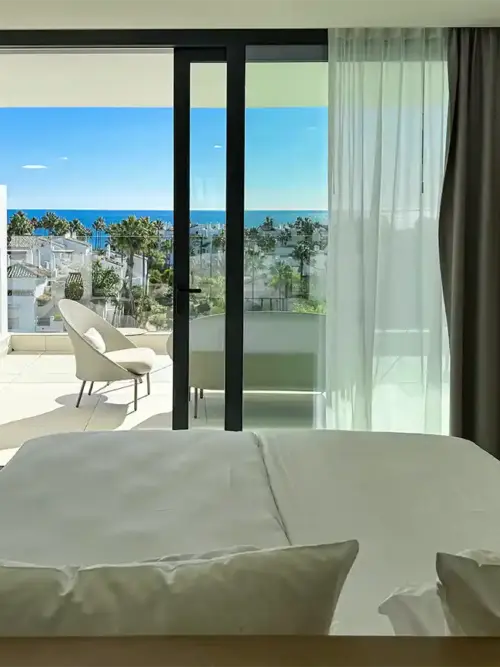 Ein helles Hotelzimmer mit einem Bett, das auf einen Balkon mit Blick auf Palmen und das Meer unter strahlend blauem Himmel ausgerichtet ist.