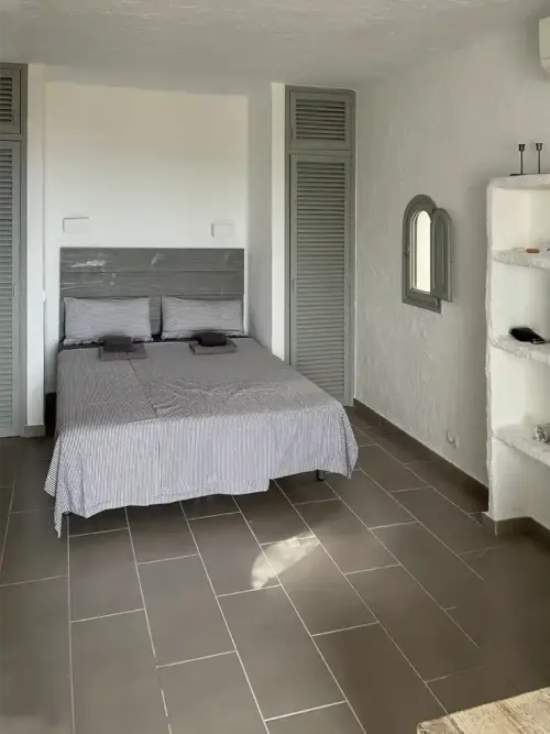 Minimalistisches Schlafzimmer mit einem Doppelbett, Einbauregalen, Fliesenboden und einem kleinen Bogenfenster.