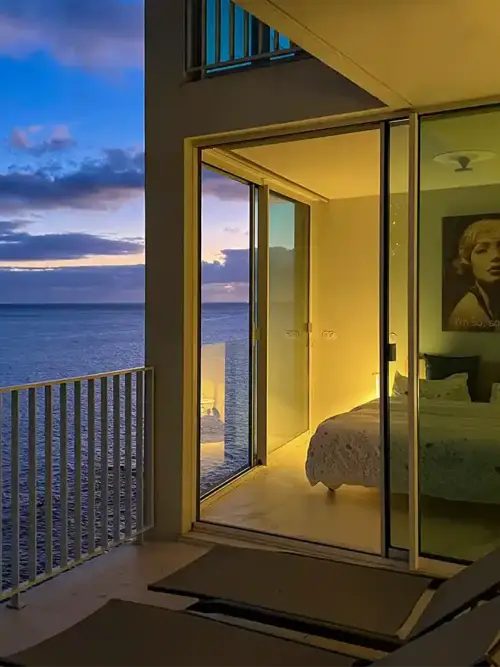 Ein modernes Schlafzimmer mit Glaswänden bietet einen Blick auf das Meer bei Sonnenuntergang, mit gemütlicher Beleuchtung im Inneren und Stühlen auf dem Balkon.