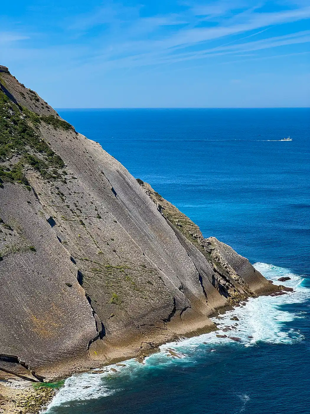 Eine felsige Steilküste trifft auf den blauen Ozean unter einem strahlenden Himmel, mit einem entfernten Boot am Horizont und den Wellen darunter.