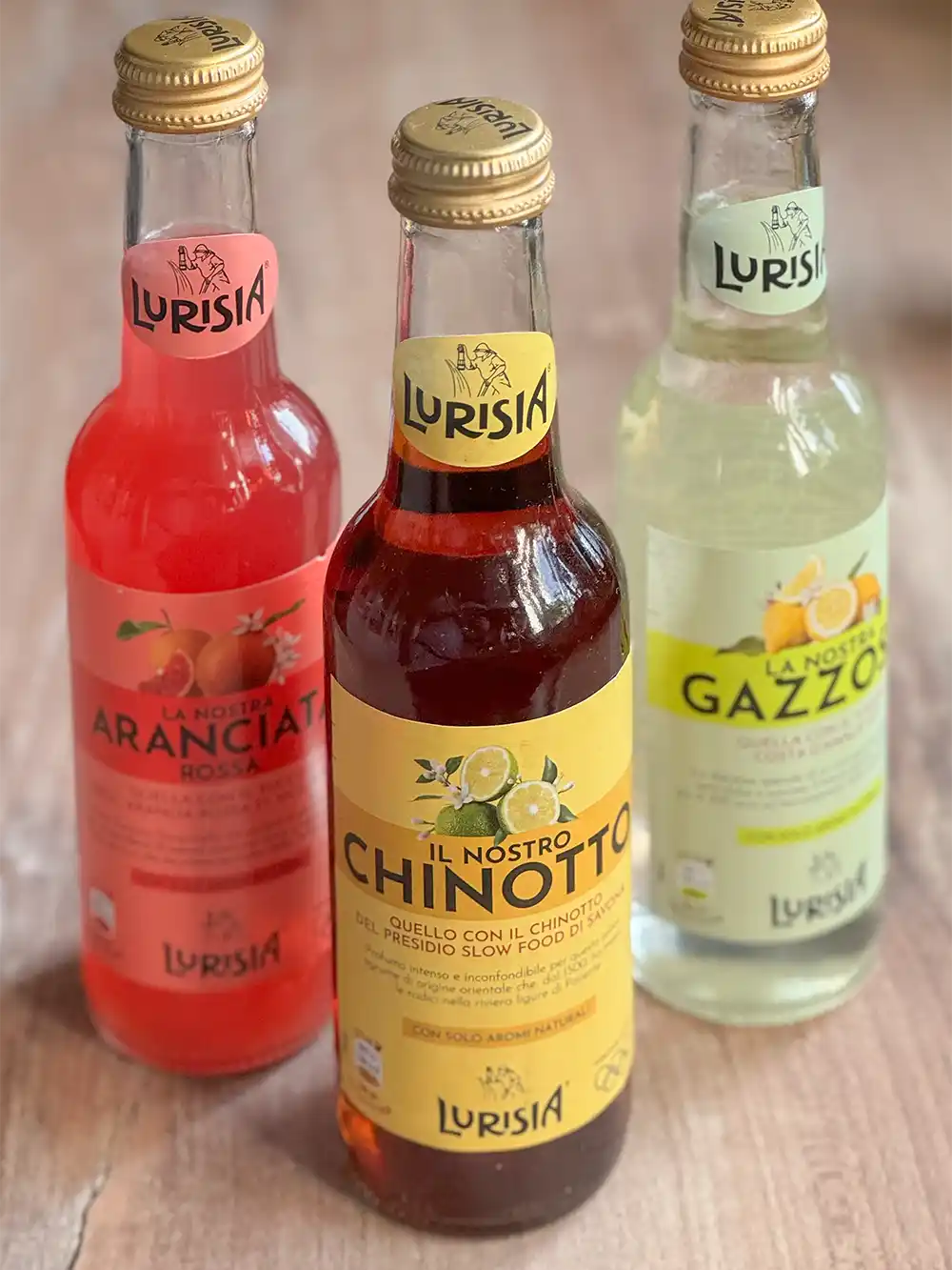 Drei Lurisia-Sodaflaschen - Aranciata, Chinotto und Gazzosa - auf einer Holzfläche.