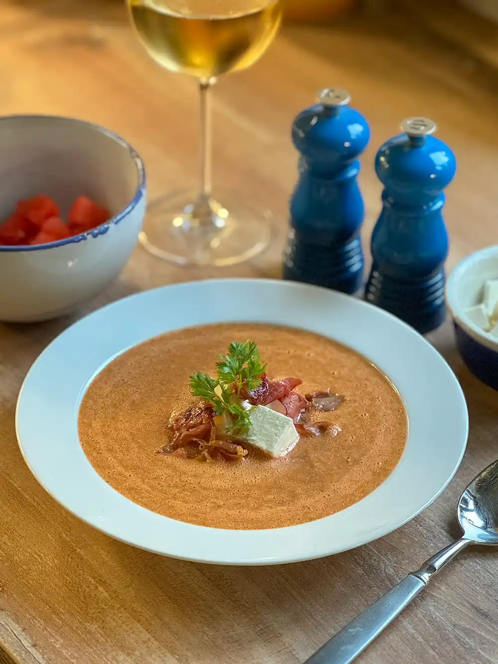 Eine Schüssel cremiger Suppe mit Kräutern, Speck und Käse, dazu Wein, Gewürze und Beilagen.