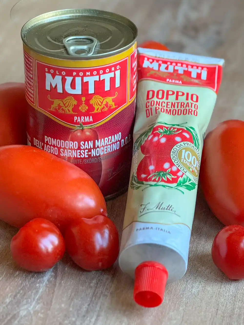 Eine Dose und eine Tube Mutti-Tomatenprodukte mit frischen Tomaten auf einer Holzunterlage.