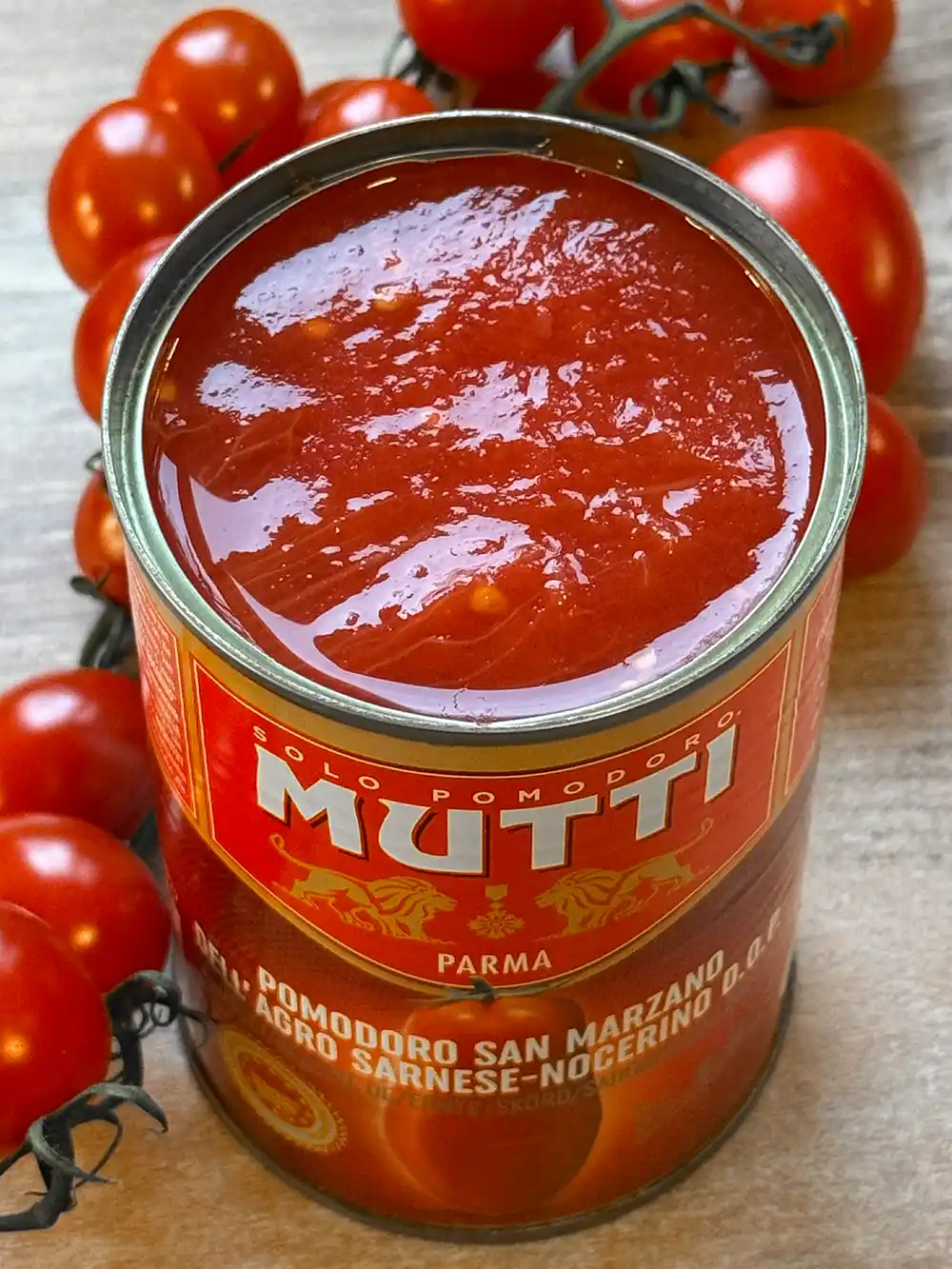 Offene Dose Mutti-Tomaten, umgeben von frischen Tomaten auf einer Holzunterlage.