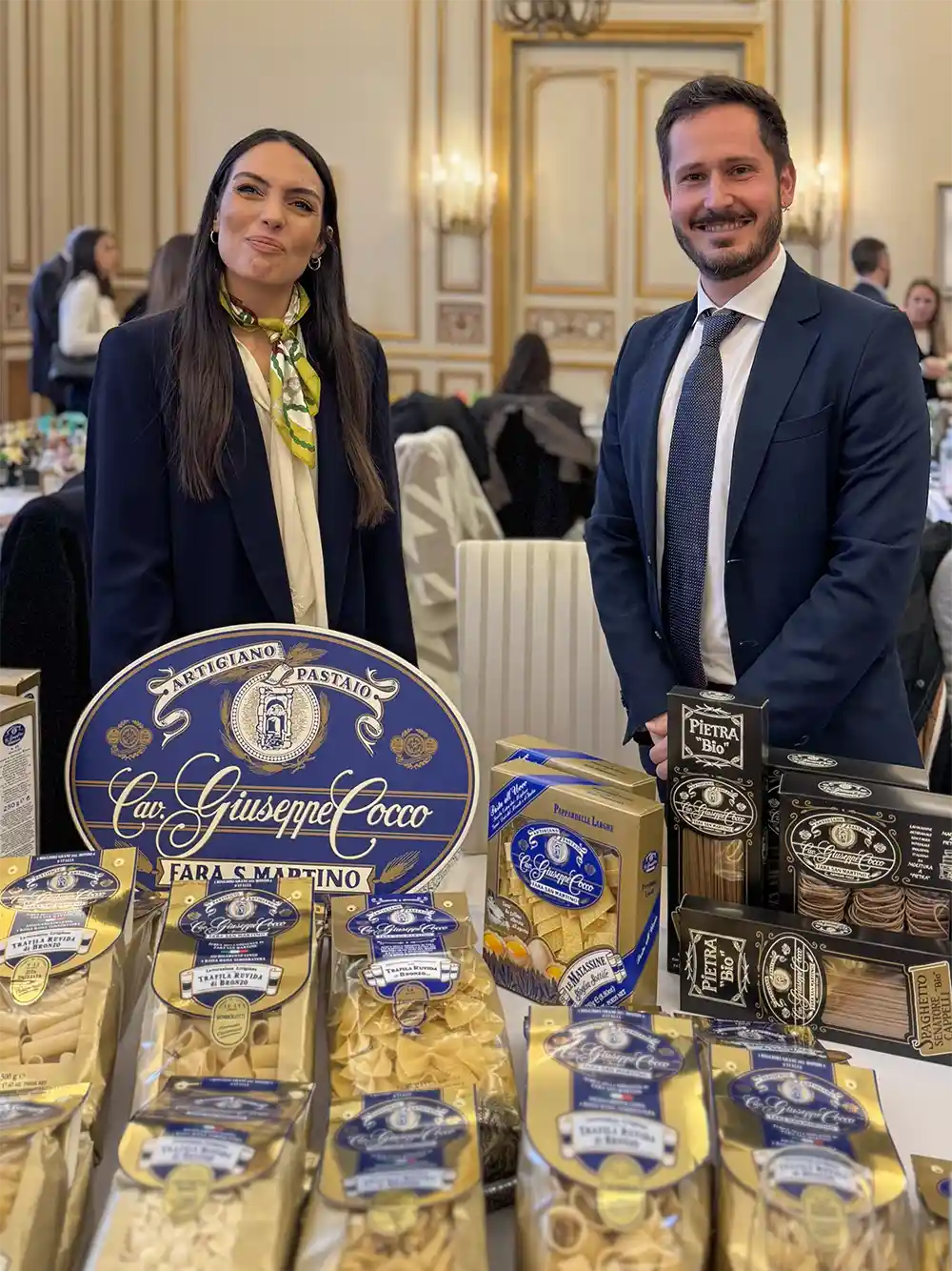 Zwei Personen stehen hinter einem Tisch, auf dem verschiedene italienische Nudelprodukte in einem formellen, eleganten Raum ausgestellt sind.