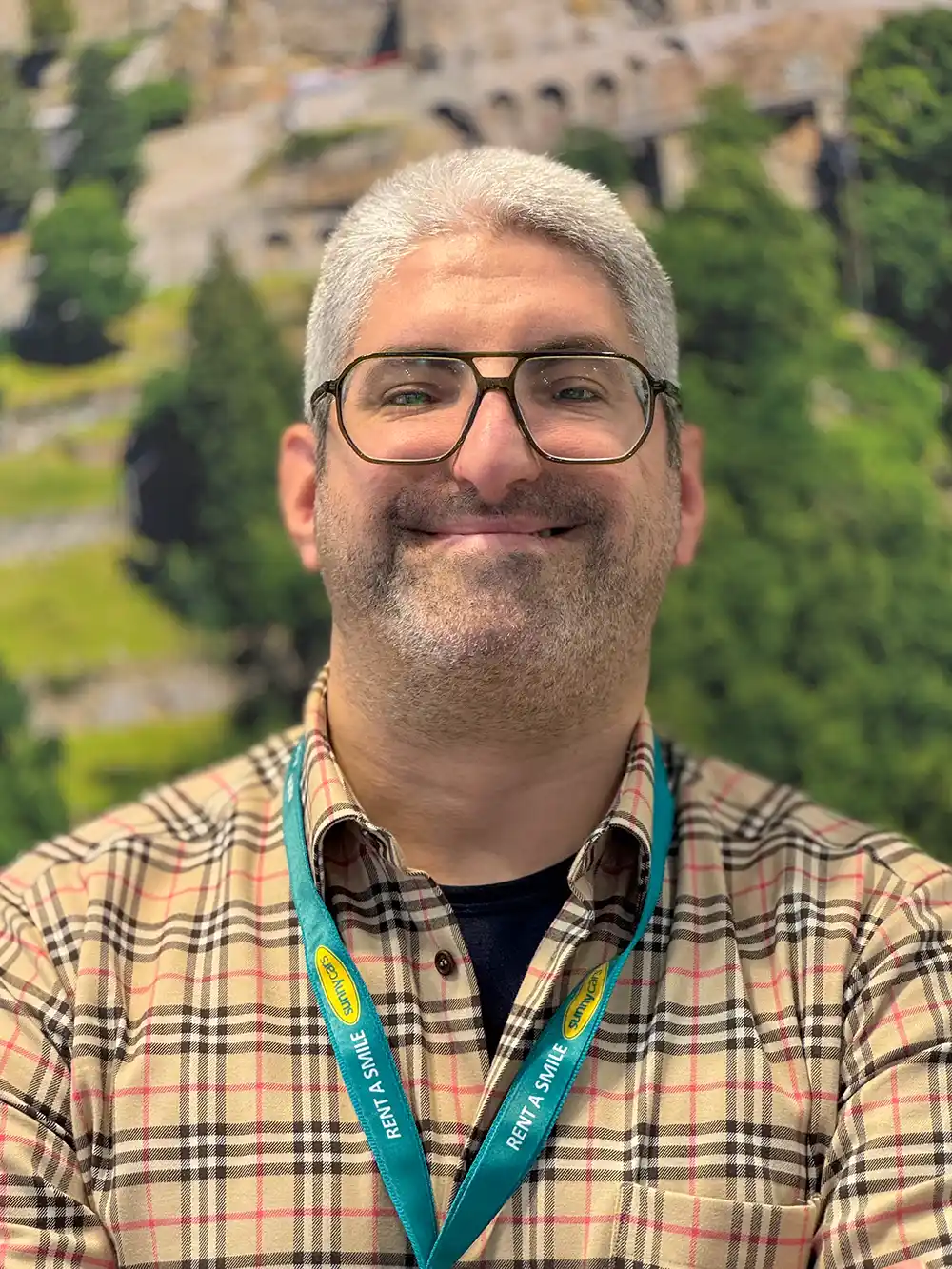 Lächelnder Mann mit Brille, der ein kariertes Hemd und ein grünes Schlüsselband trägt und vor einem Naturhintergrund steht.