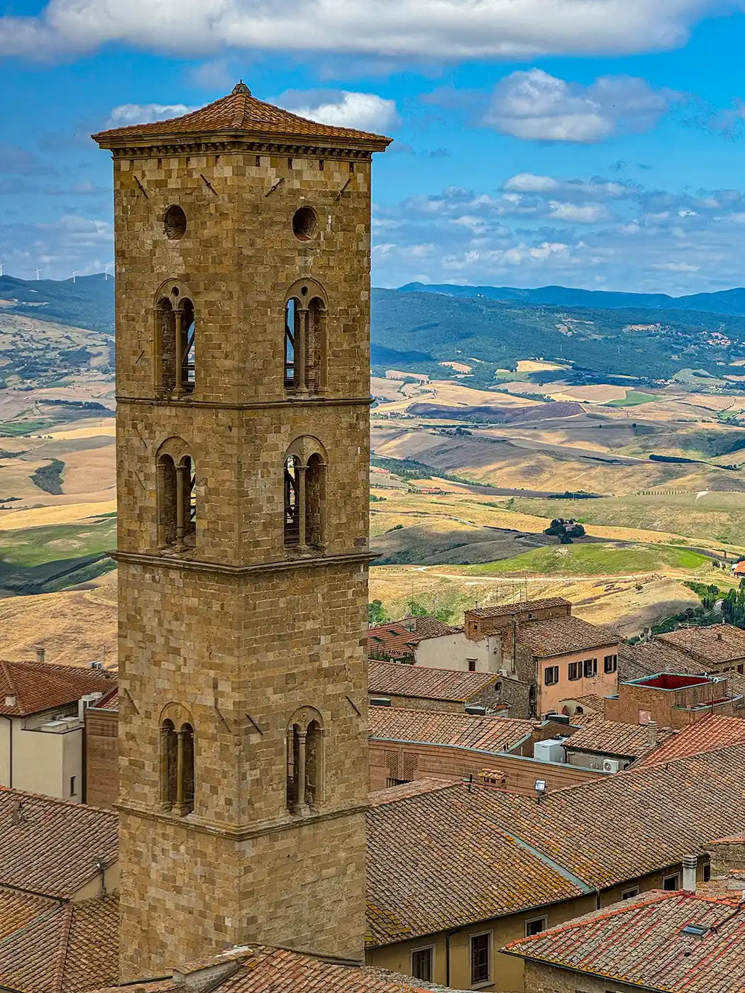Ein hoher steinerner Glockenturm erhebt sich über Terrakotta-Dächern, mit sanften Hügeln und blauem Himmel im Hintergrund.