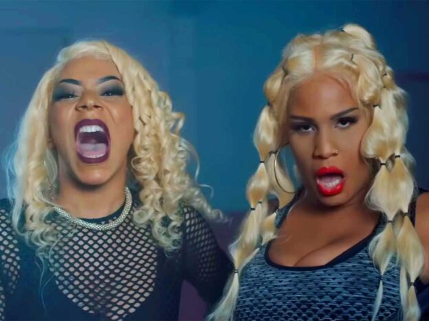 Zwei Personen mit langen blonden Haaren und auffälligem Make-up posieren, wobei die eine schreit und die andere in die Kamera schaut.