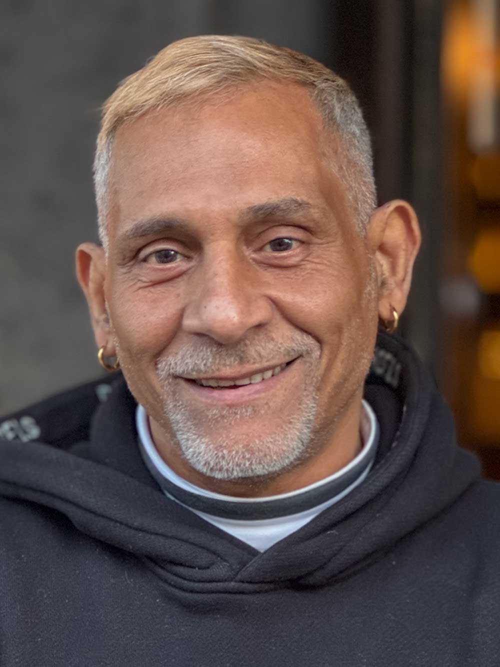 Lächelnder Mann mit kurzen blonden Haaren, grauem Spitzbart, schwarzer Kapuzenjacke und goldenen Ohrringen.