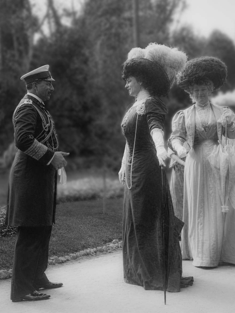 Ein Mann in Uniform begrüßt zwei Frauen in aufwendigen Kleidern und großen Hüten in einem Garten.