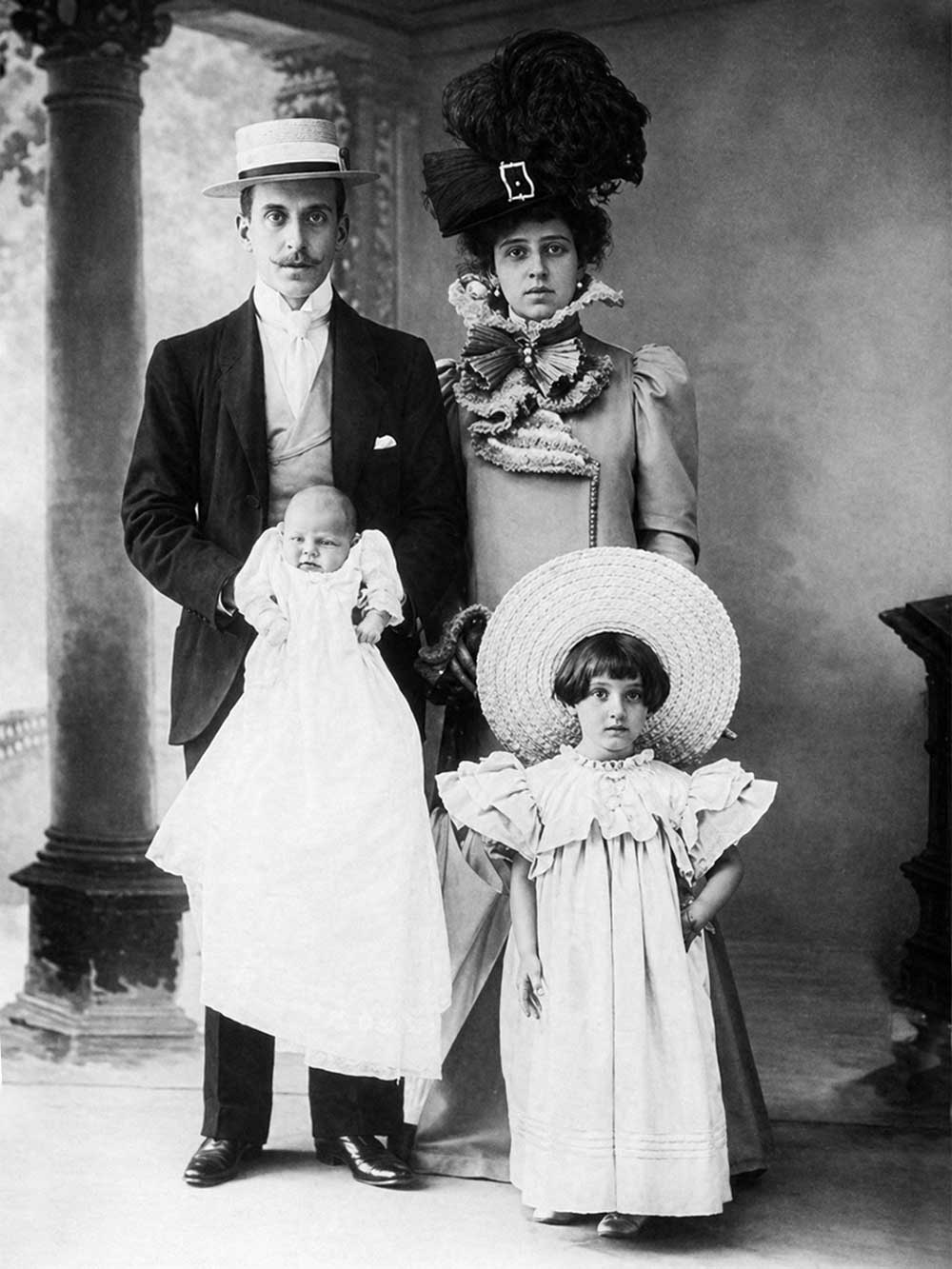 Eine Familie aus dem viktorianischen Zeitalter posiert für ein Porträt: Vater, Mutter mit großem Hut, junges Mädchen und ein Baby im Kleidchen.