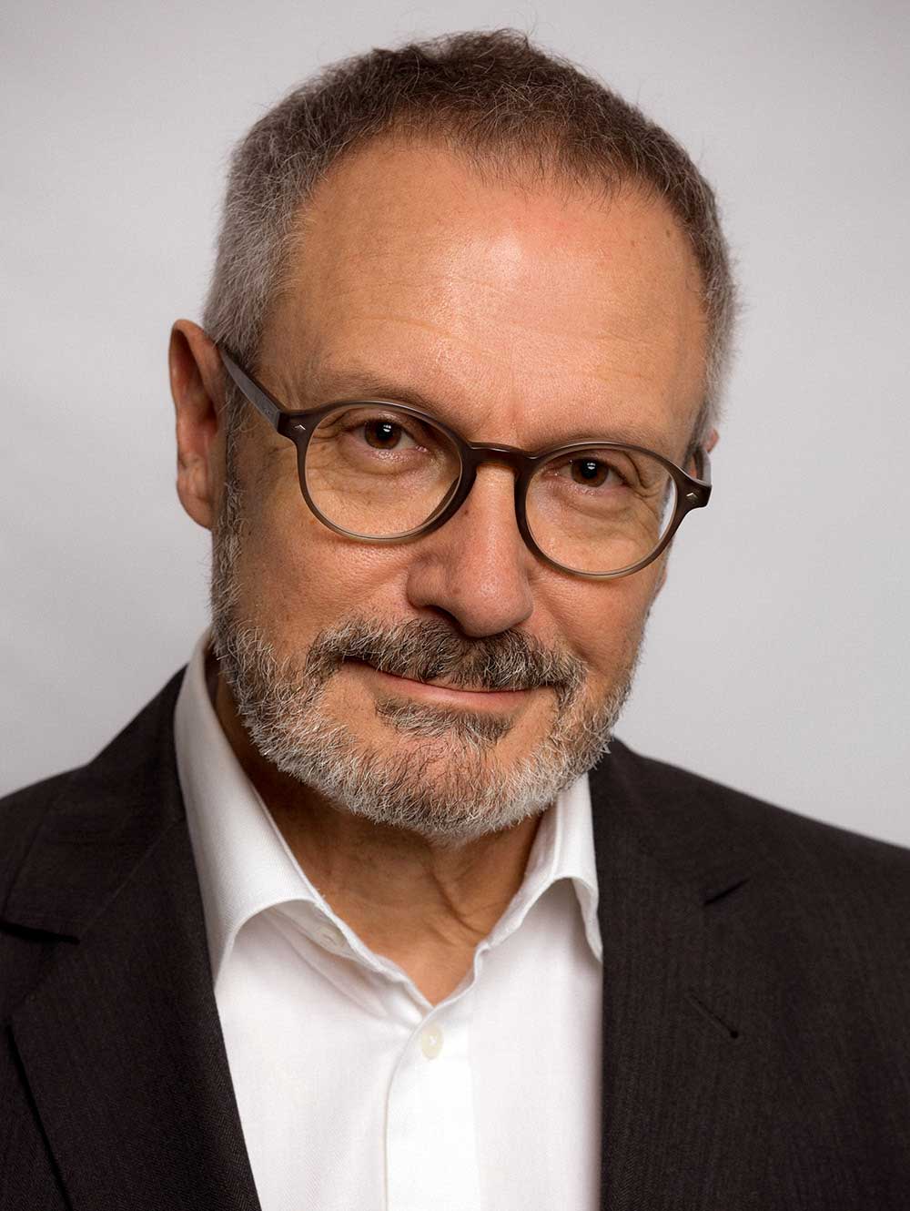 Ein Mann mittleren Alters mit Brille, kurzen grauen Haaren und einem Bart, der einen dunklen Anzug und ein weißes Hemd trägt.