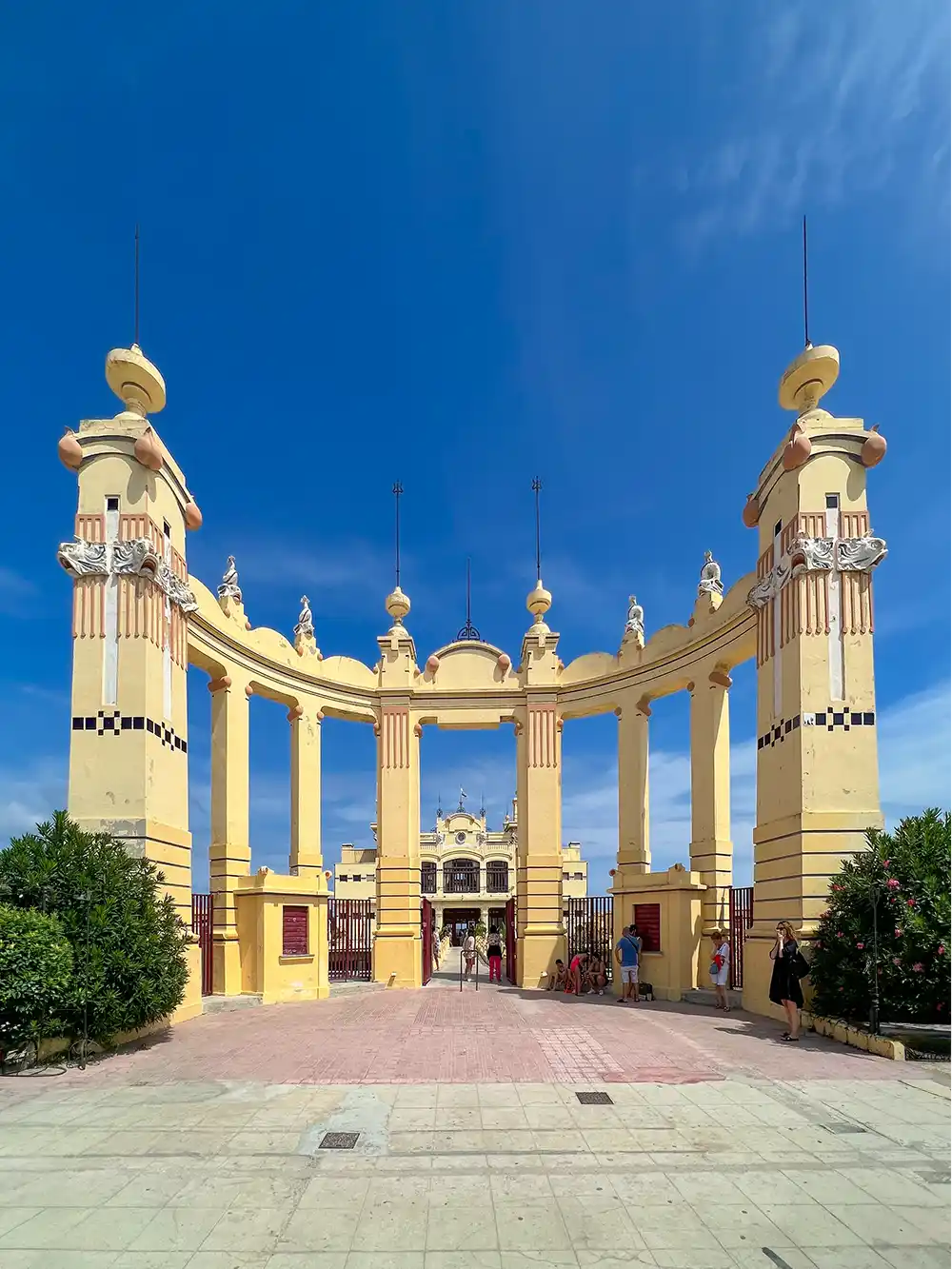 Gelbes neoklassizistisches Eingangstor mit Säulen unter strahlend blauem Himmel, Menschen gehen in der Nähe spazieren.