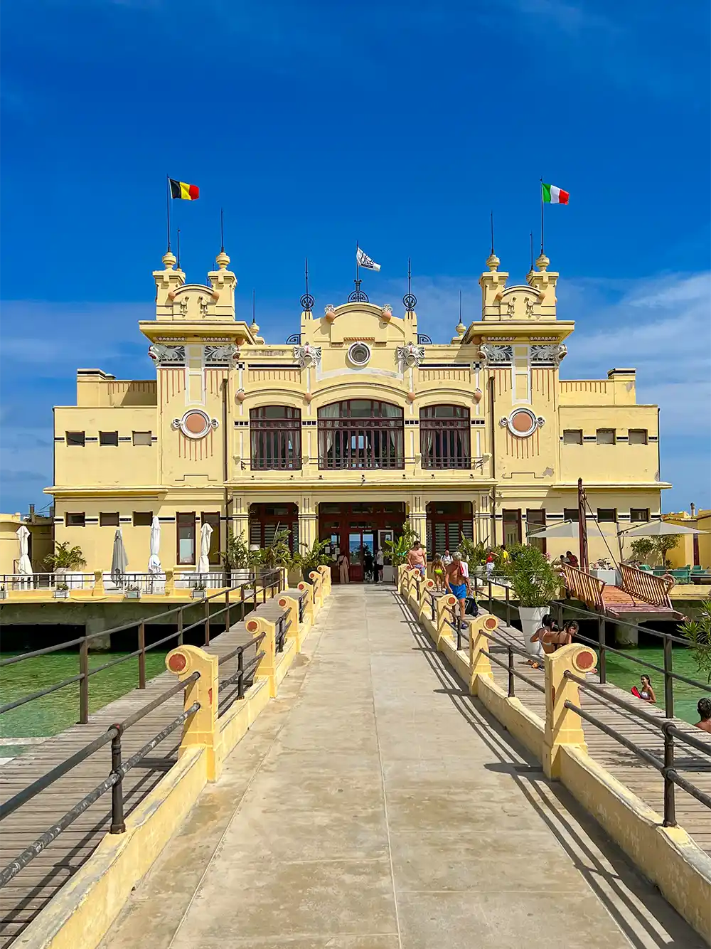 Ein gelbes historisches Gebäude mit Fahnen, betrachtet von einem Gehweg aus unter einem strahlend blauen Himmel.
