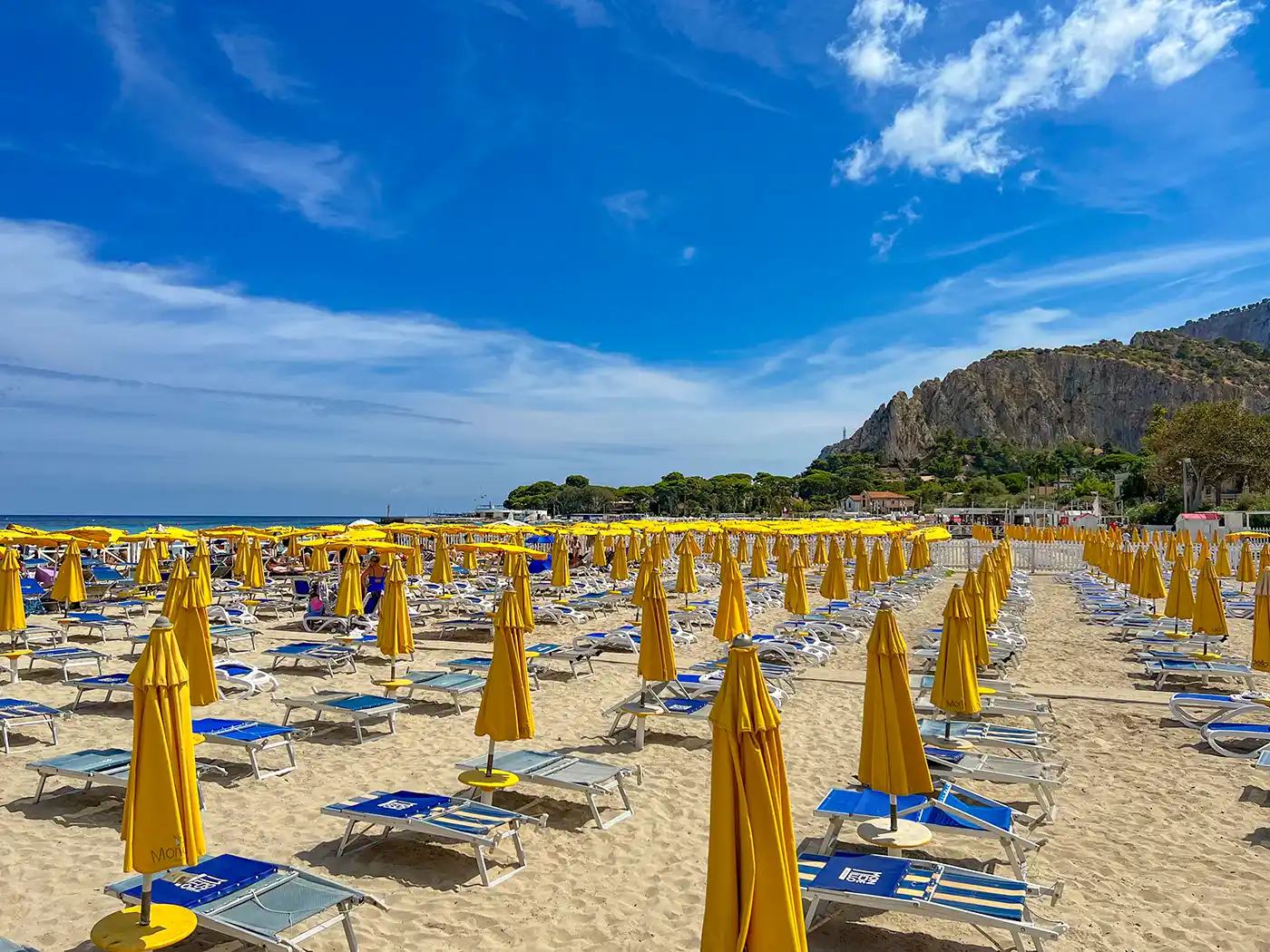 Reihen von gelben Sonnenschirmen und blauen Liegestühlen an einem Sandstrand mit Klippen und blauem Himmel im Hintergrund.