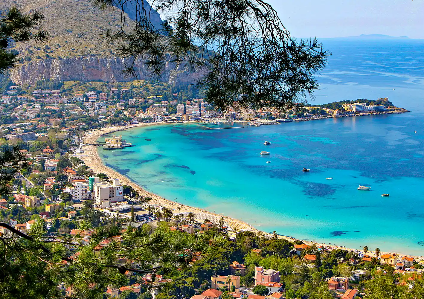 Luftaufnahme einer türkisfarbenen Bucht, eines Sandstrands und einer Küstenstadt mit Hügeln und Bäumen im Hintergrund.