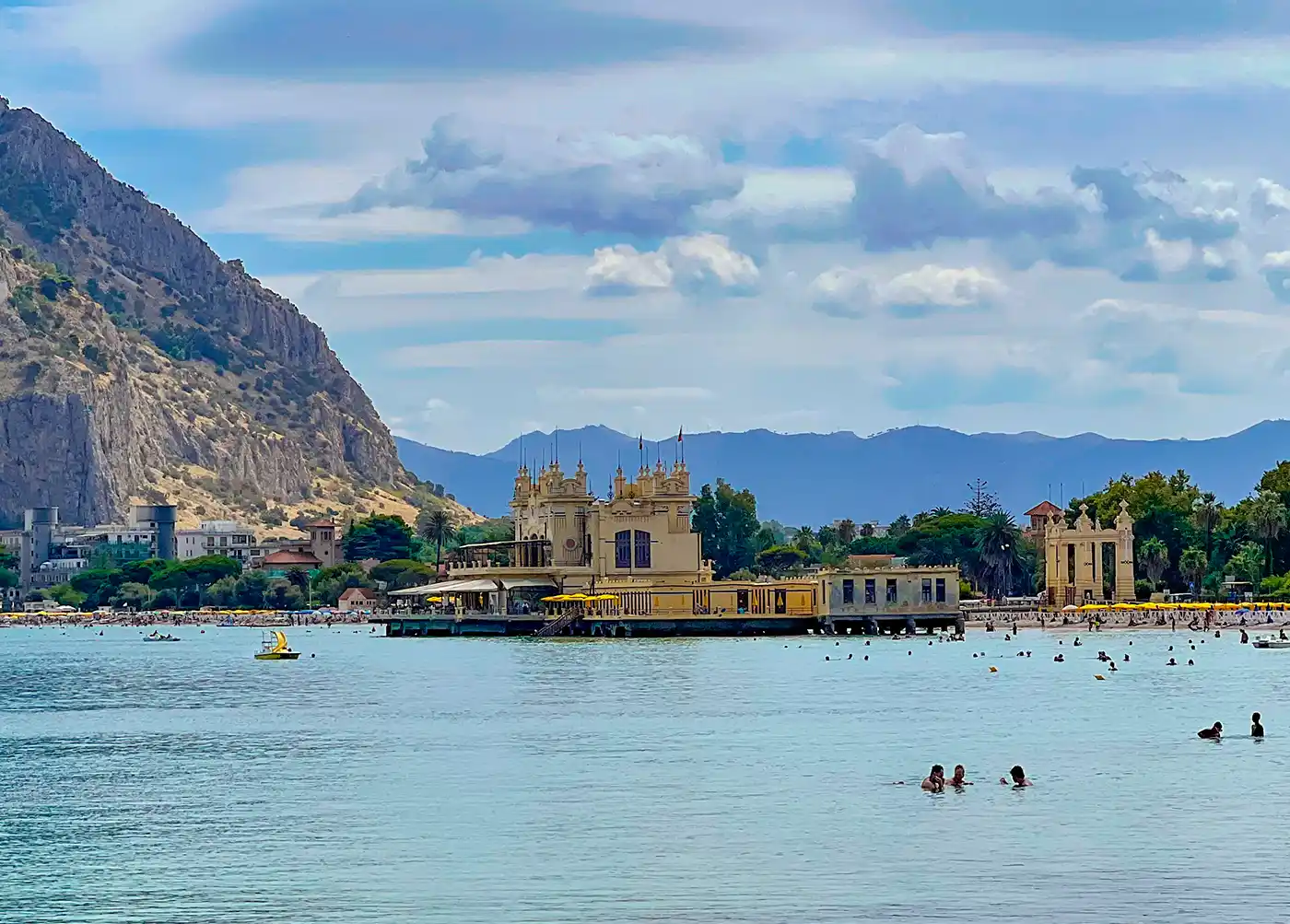Eine Strandszene mit Schwimmern, einem historischen Gebäude am Wasser und Bergen im Hintergrund.