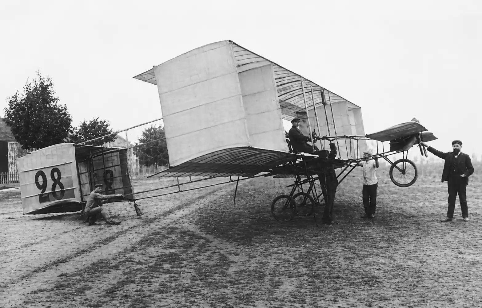 Frühes Oldtimer-Flugzeug auf einem grasbewachsenen Feld mit einem sitzenden Piloten und mehreren in der Nähe stehenden Personen.
