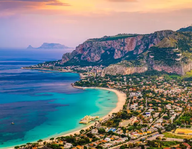 Türkisfarbene Bucht mit Sandstrand, felsigen Klippen und einer Küstenstadt bei Sonnenuntergang unter einem bunten Himmel.