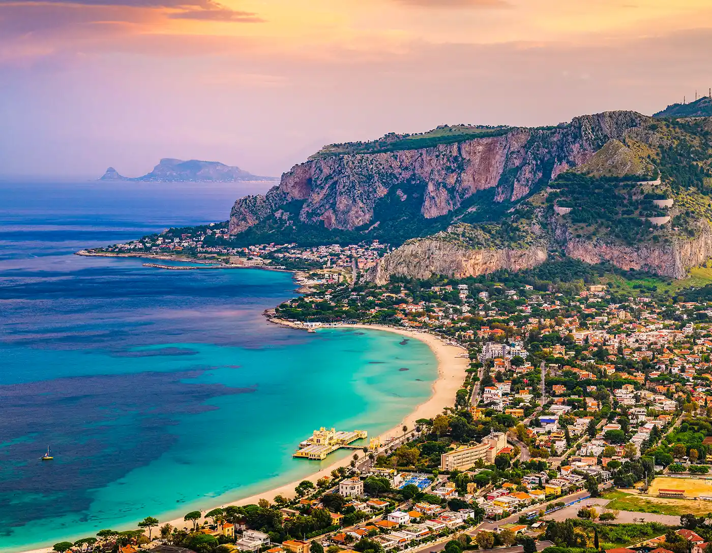Türkisfarbene Bucht mit Sandstrand, felsigen Klippen und einer Küstenstadt bei Sonnenuntergang unter einem bunten Himmel.