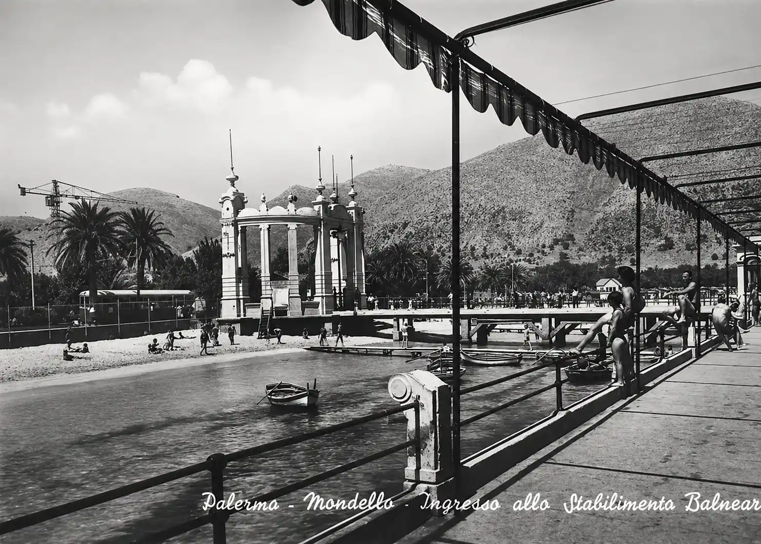 Menschen spazieren entlang einer Strandpromenade in Mondello, Palermo, mit Bergen und einem Pavillon im Hintergrund.