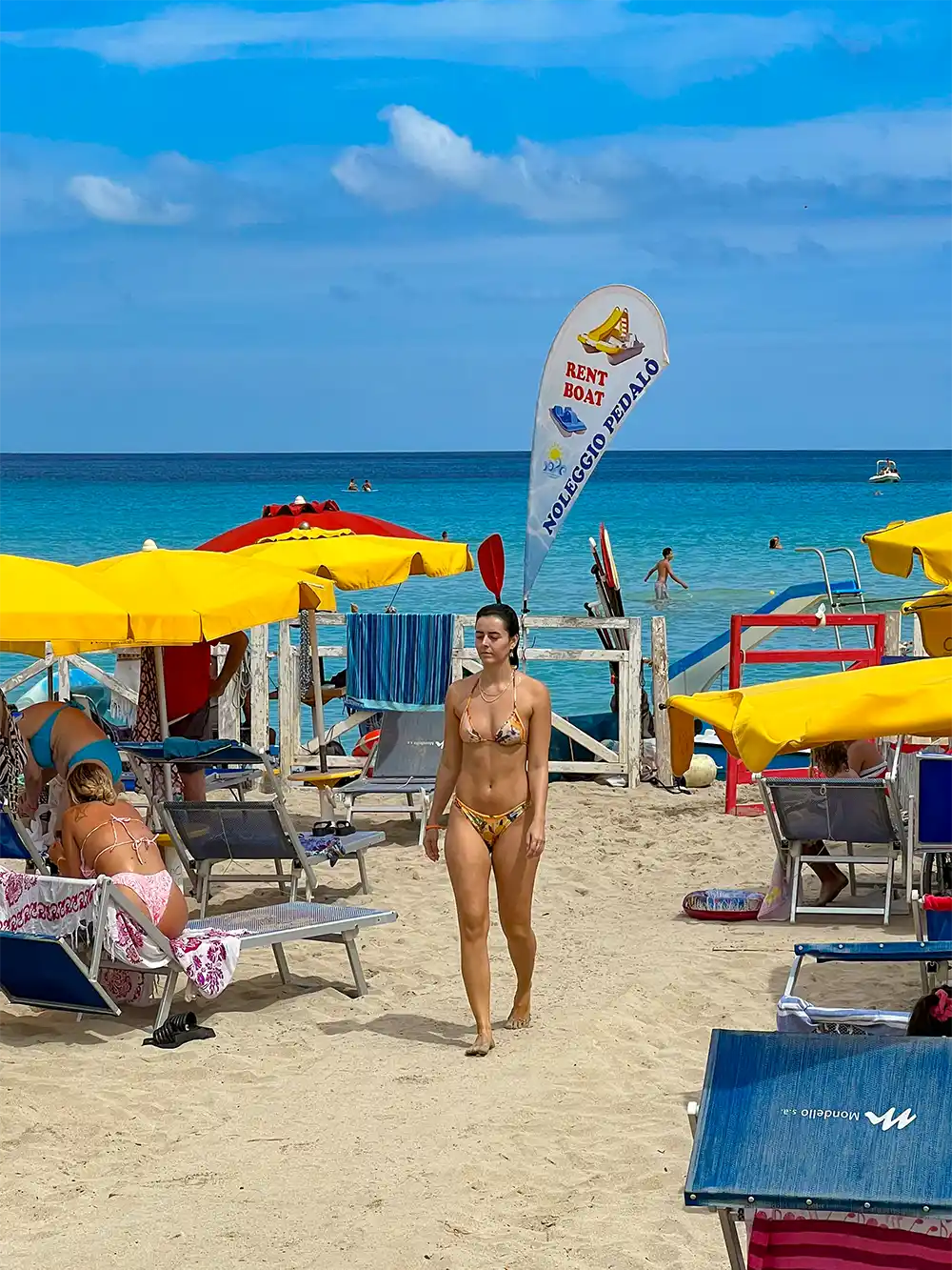 Frau im Bikini spaziert an einem belebten Strand mit gelben Sonnenschirmen, Liegestühlen und einem "Rent Boat"-Schild.