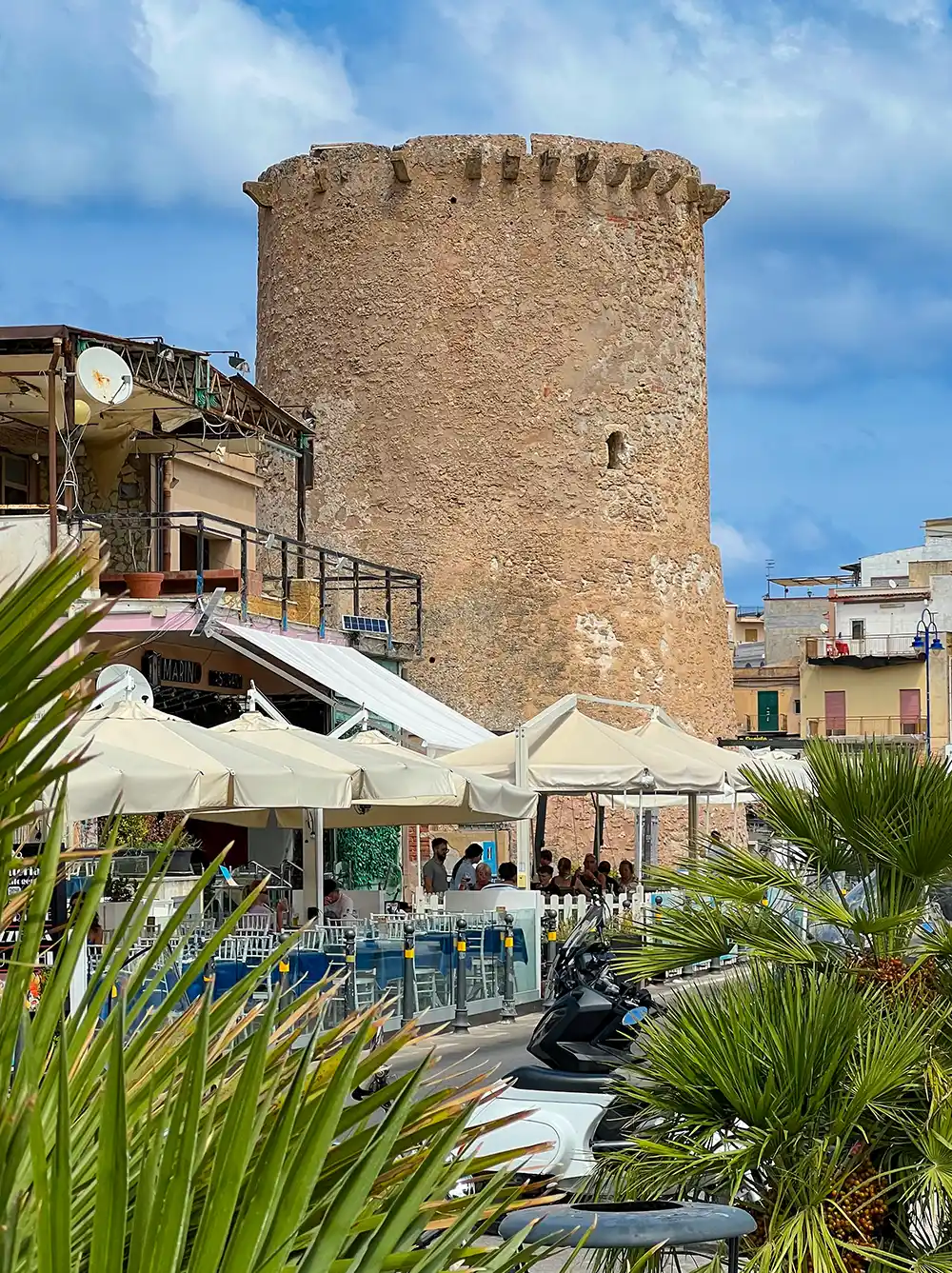 Alter Steinturm hinter einem Straßencafé mit weißen Sonnenschirmen und Palmen unter einem teilweise bewölkten Himmel.