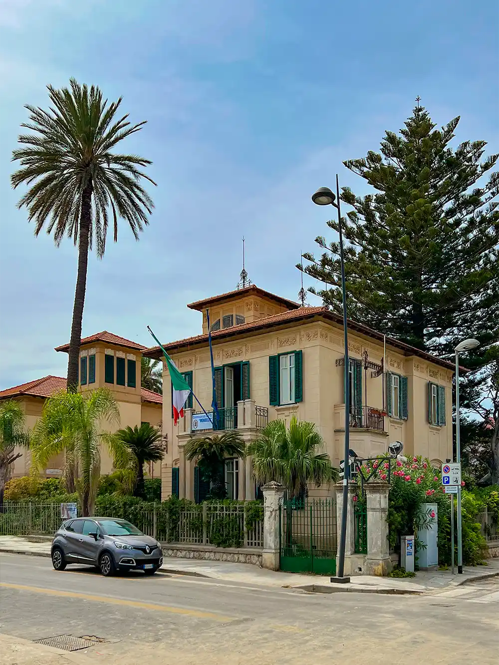 Eine Villa mit Palmen, italienischer Flagge und einem geparkten Auto an einem sonnigen Tag.