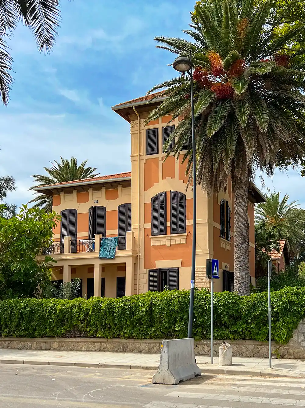 Eine hellbraune und orangefarbene Villa mit dunklen Fensterläden, Palmen und einem Balkon steht hinter einer grünen Hecke.