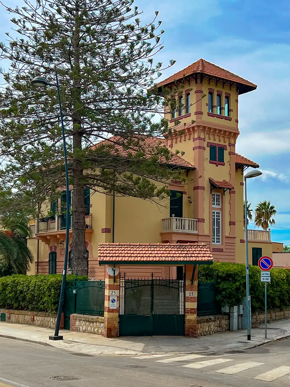 Eine gelbe Villa mit einem Turm, roten Dachziegeln und grünen Fensterläden, hinter einem Tor an einer sonnigen Straßenecke.