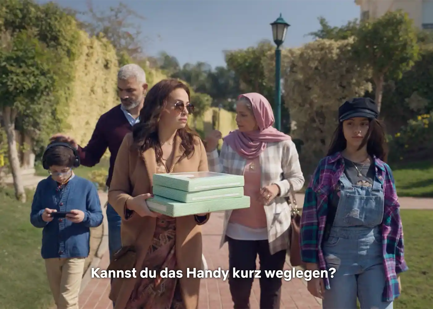 Eine Familie geht nach draußen; eine Frau trägt Kisten. Untertitel: Kannst du das Handy kurz weglegen?.