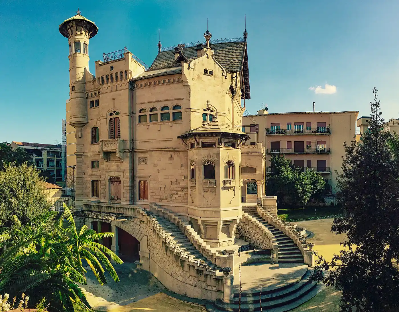 Ein großartiges historisches Herrenhaus mit einem Turm, Bogenfenstern und geschwungenen Steintreppen unter einem klaren blauen Himmel.