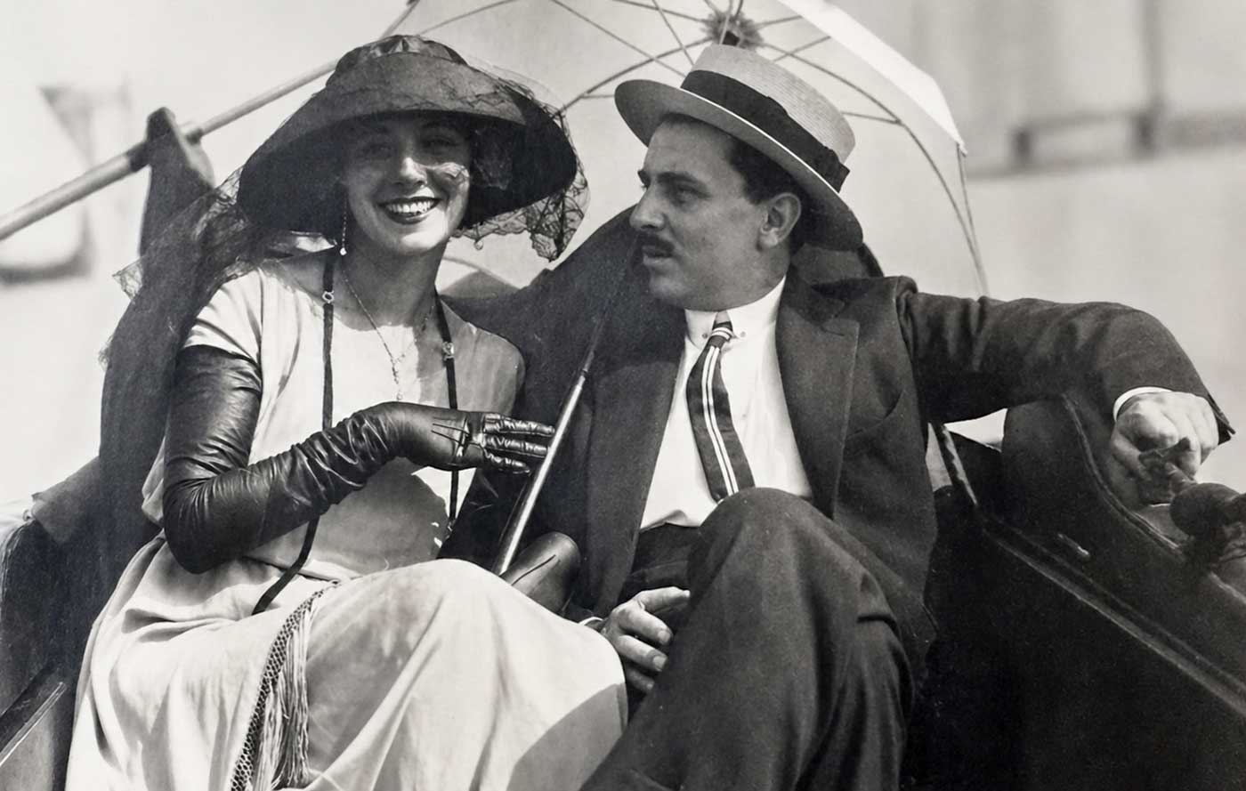 Eine Frau mit Hut und Handschuhen lächelt neben einem Mann in Anzug und Hut, der unter einem Sonnenschirm sitzt.