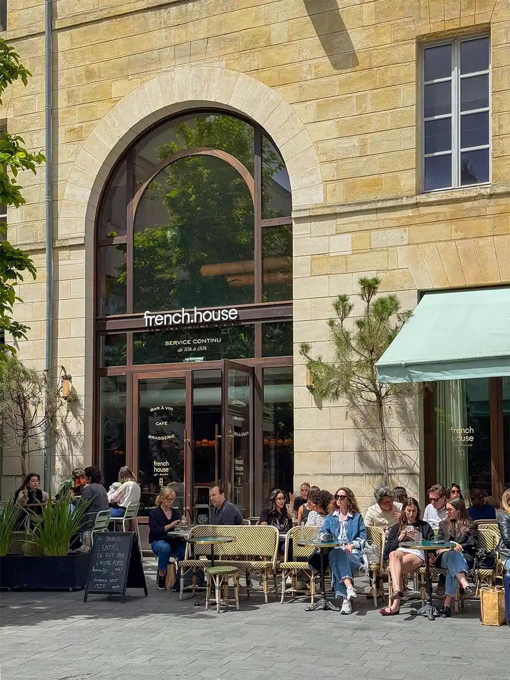 An einem sonnigen Tag sitzen Menschen an Tischen im Freien vor einem Café namens french.house.